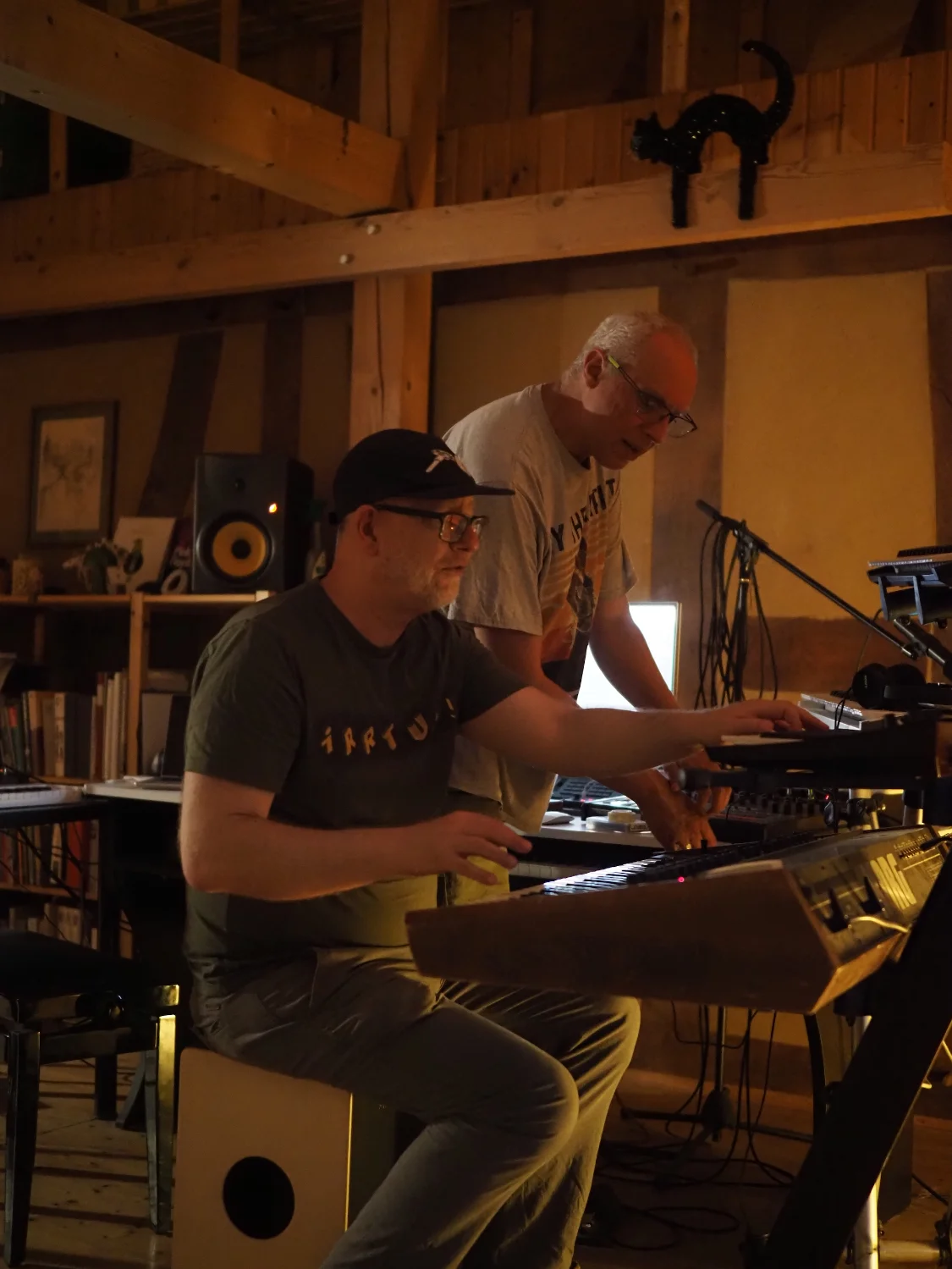 Photo by Frau K.: Herr Irrtum! sitting on the left, wearing a Basspistol cap and an Irrtum! shirt, tinkering with two synthesizers, while Stephan/Schall und Stille is standing behind him, wisely advising and gently manipulating synth settings.
Both are very focused.
The studio where all this takes place is bathed in warm evening light.